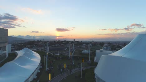 Luftaufnahme,-Drohne,-Blick-Auf-Den-Sonnenuntergang-Vom-Einkaufszentrum,-Blick-Auf-Den-Parkplatz-Mit-Vielen-Autos,-Windkraft,-Festzelt