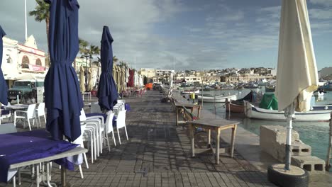 Riviera-Von-Marsaxlokk-Mit-Geschlossenen-Cafés,-Sonnenschirmen-Und-Booten,-Die-An-Sonnigen-Tagen-In-Der-Bucht-Schaukeln