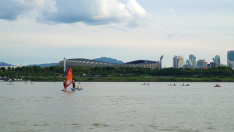 Windsurfer-And-Paddle-Boarding-In-Han-River-With-Jamsil-Olympic-Stadium-At-Background-In-Seoul,-South-Korea