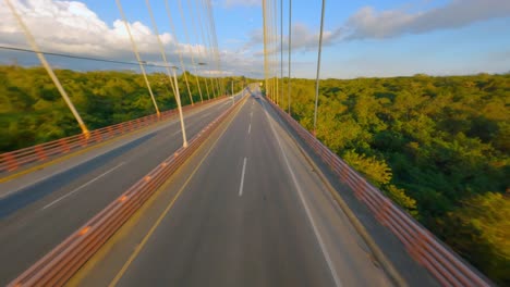 Drone-Low-Overhead-View-of-Traffic-Driving-on-Mauricio-Baez-Suspension-Bridge-Road-in-Early-Morning-Sunlight,-FPV