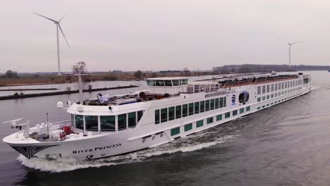 Aerial-View-Of-River-Cruise-Ship-Navigating-Along-Oude-Maas-Going-Past-Still-Wind-Turbines-In-Barendrecht