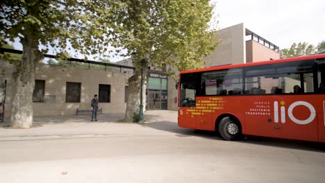 Red-Bus-Regional-Occitanie-Transport-Lio-arriving-at-bus-stop-with-people-waiting,-Pan-left-follow-shot