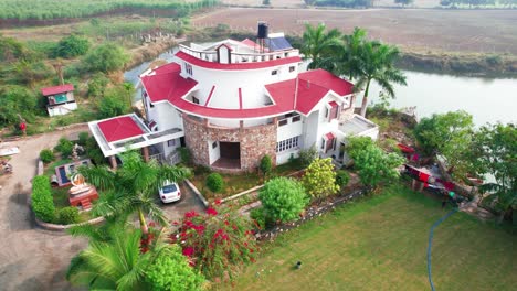 Video-traveling-sideways-over-palm-trees-and-red-and-green-vegetation-at-the-vacation-home-for-relaxation-by-the-lake,-pool-and-parking-lot-in-Vadodara,-India