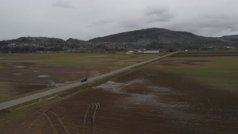 cars-driving-on-country-farm-road-in-Abbotsford-BC-Wide-Aerial-fixed-position-scenic-view-of-highway-wet-fields-grass-crops-wheat-vegetables-in-valley-surrounded-by-mountains