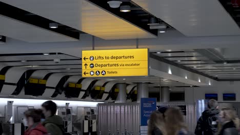 Gelbe-Wegweiser-Am-Terminal-3-Des-Internationalen-Flughafens-Heathrow,-Vorbeikommende-Passagiere,-Die-Ihren-Geschäften-Nachgehen,-Auf-Dem-Weg-Zum-Abflugterminal,-London,-England
