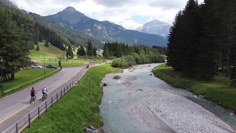 Val-di-Fassa-at-Trentino,-Dolomites,-Italy---Aerial-Drone-View-of-Cycling-Tourists,-Blue-River-and-Green-Mountain-Valley