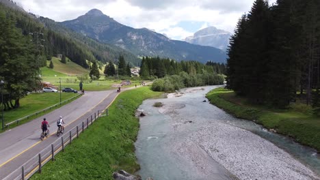 Val-Di-Fassa-En-Trentino,-Dolomitas,-Italia---Vista-Aérea-De-Drones-De-Ciclistas,-Río-Azul-Y-Valle-De-Montaña-Verde