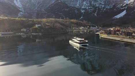 Visión-Del-Catamarán-De-Pasajeros-De-Los-Fiordos-Que-Se-Acercan-Al-Puerto-De-Flam-Durante-El-Hermoso-Amanecer-De-La-Mañana---Seguimiento-Aéreo-Y-Cruce-Detrás-Del-Barco-Con-Rayos-De-Sol-Y-Reflejos-Que-Golpean-El-Catamarán-Y-El-Mar