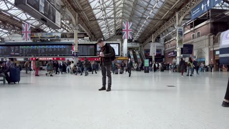 Innenansicht-Der-Londoner-Victoria-Station-Mit-Einem-Erwachsenen-Mann,-Der-Sein-Telefon-überprüft,-Und-Pendlern,-Die-Im-Hintergrund-Laufen