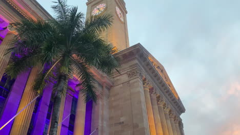 Orangefarbener-Sonnenuntergangsschwenkblick-Auf-Den-Uhrturm-Des-Rathauses-Von-Brisbane-Mit-Violetten-Lichtern,-Die-Die-Palmen-Erhellen