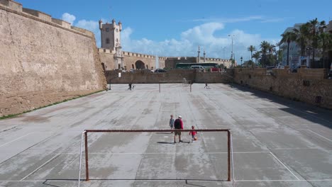Children-play-at-a-football-playground-near-the-Puerta-de-Tierra,-and-ancient-city-wall-in-Cadiz,-Andalusia,-southern-Spain