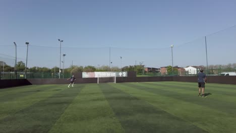 Two-Males-Kicking-Football-In-The-Air-To-Each-Other-As-Warm-Up-On-Soccer-Pitch-At-Goals-On-24-April-2022