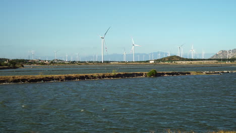 Static-low-POV-of-wind-turbines-against-blue-sky,-river-in-foreground,-Vietnam