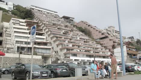 Hillside-Commercial-Structure-And-Apartment-Buildings-Beside-A-Public-Road-With-Parked-Cars-And-People-Idling-On-A-Bench-In-Renaca,-Vina-Del-Mar,-Chile