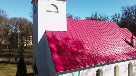 Revealing-aerial-view-of-Lielvarde-Lutheran-church-at-the-bank-of-Daugava-river,-white-church-with-red-roof,-leafless-trees,-sunny-spring-day,-medium-ascending-shot