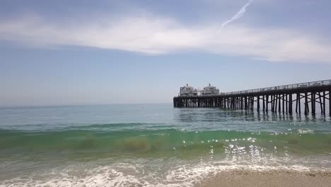 Wave-breaks-on-the-beach,-flying-along-the-landmark-of-Malibu-Pier