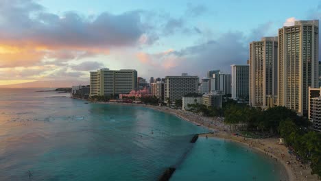 Hawaiian-Beach-Surfers-and-Tourists-Near-Beachfront-Resort-Hotel-Buildings-Swimming-and-Relaxing-in-Sand-On-Waikiki-Beach-In-Honolulu,-Hawaii-At-Sunset