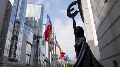 Filmische-Ansicht-Der-Göttin-Europa,-Die-Das-Euro-Symbol-Vor-EU-Flaggen-Vor-Dem-Paul-Henri-Spaak-Gebäude-Im-Europäischen-Parlament-Hält