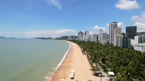 aerial-drone-flying-center-with-the-tropical-white-sand-beach-on-a-sunny-day-full-of-coconut-trees-and-tall-beachside-hotels-and-beautiful-turquoise-ocean-on-the-left-side-located-in-Nha-Trang-Vietnam