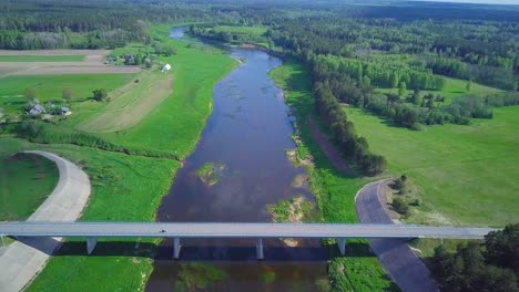 Aerial-view-of-a-Venta-river-on-a-sunny-summer-day,-lush-green-trees-and-meadows,-beautiful-rural-landscape,-wide-angle-drone-shot-moving-forward-over-the-white-concrete-bridge