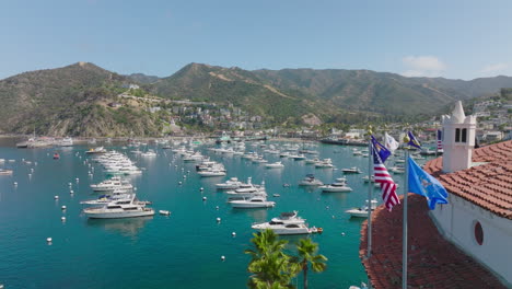 Flying-Over-Catalina-Island,-Drone-Footage-Approaching-Sparkling-Blue-Harbor-filled-with-Boats-and-Mountains-in-Background