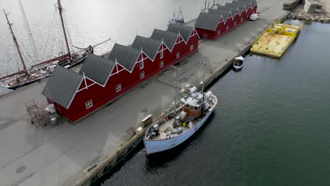 Aerial-view,-old-wooden-ship-is-moored-in-the-port-at-the-quay,-on-the-pier-stand-the-wooden-fishermans-warehouses