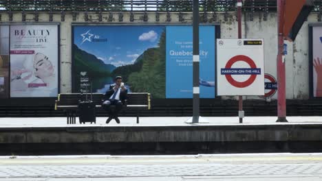 UK-Asian-Businessman-Seated-On-Station-Platform-Checking-His-Phone-On-The-London-Underground-In-London,-27-May-2022