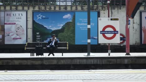 Reino-Unido-Empresario-Asiático-Sentado-En-La-Plataforma-De-La-Estación-Revisando-Su-Teléfono-En-El-Metro-De-Londres,-27-De-Mayo-De-2022