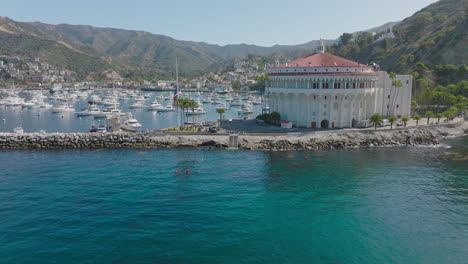 Drone-Flying-Through-Avalon-Harbor,-Over-Boats-on-Bright-Turquoise-Blue-Waters-of-Catalina-Island,-Avalon-Resorts-and-Homes-Nestled-In-Mountain