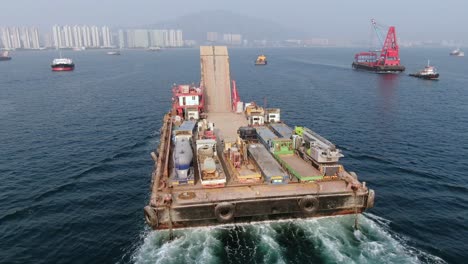 Barge-loaded-with-Concrete-mixer-trucks-pulled-to-port-by-a-Tugboat-in-Hong-Kong-bay,-Aerial-view