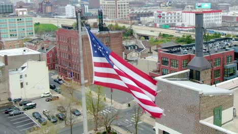 Aerial-view-around-the-American-flag,-waving-in-the-wind,-with-city-background---orbit,-drone-shot