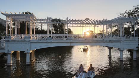 Aerial-dolly-out-of-people-sailing-in-boats-under-white-bridge-in-Rosedal-gardens-at-golden-hour,-Buenos-Aires