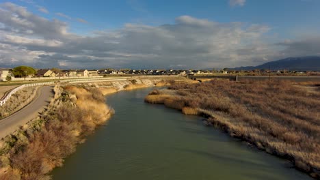 Aerial-footage-of-a-river-flowing-towards-the-distant-mountains-and-clouds-along-side-a-paved-biking-trail