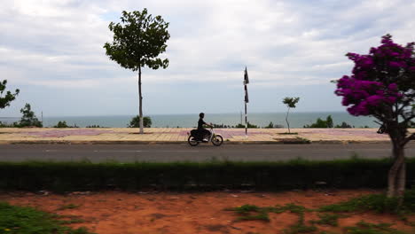 Dolly-shot-of-a-young-girl-riding-her-scooter-on-an-empty-seaside-road-in-South-East-Asia