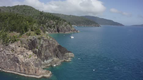 Aerial-flight-past-shattered-rocky-cliffs-to-catamaran-sailboat-in-bay