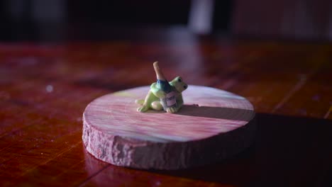 Cute-Frog-Figurine-Made-Of-Ceramic-Holding-Miniature-Bottle-Of-Champagne-On-A-Wooden-Base,-Illuminated-By-Glowing-Projector-Lights