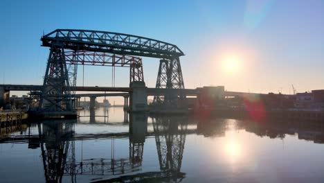 Low-level-aerial-push-in-shot-of-ferry-bridge-Nicolas-Avellaneda-over-the-Matanza-River-during-sunrise-with-sun-flare-reflection
