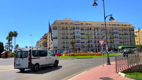 Spanish-street-with-cars-driving-in-city-Estepona-with-palm-trees,-mountain-and-colorful-flowers,-Paseo-Maritimo