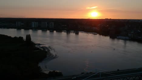 Brücke-über-Den-Fluss-Mit-Verkehr-Und-Silhouette-Der-Stadt-Bei-Sonnenuntergang,-Luftaufnahme