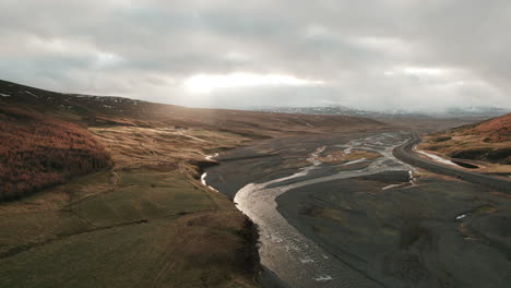 Reverse-aerial-dolly-over-beautiful-river-valley-in-Iceland,-illuminated-by-sunbeams-through-dark-clouds