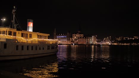 Vista-Estática-De-Un-Barco-De-Vapor-Bellamente-Decorado-Atracado-En-La-Orilla-Del-Río-Por-La-Noche-Con-Vista-De-La-Ciudad-Al-Fondo-En-El-Centro-De-Estocolmo,-Suecia