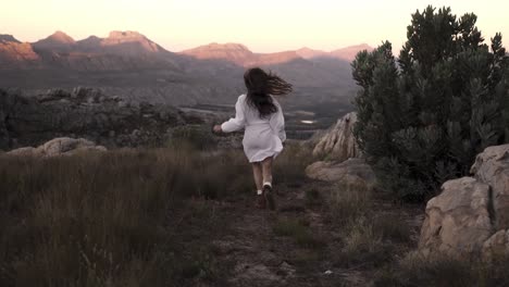 Unrecognizable-woman-running-towards-the-mountains-range-in-a-rocky-environment,-slow-motion-freedom-and-adventure-scene-seen-from-the-back