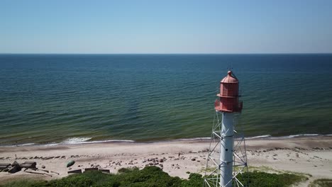 Beautiful-aerial-view-of-white-painted-steel-lighthouse-with-red-top-located-in-Pape,-Latvia-at-Baltic-sea-coastline-in-sunny-summer-day,-distant-sea,-wide-angle-drone-shot-moving-forward-slow
