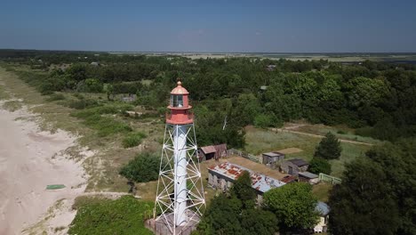 Beautiful-aerial-view-of-white-painted-steel-lighthouse-with-red-top-located-in-Pape,-Latvia-at-Baltic-sea-coastline-in-sunny-summer-day,-wide-angle-descending-drone-shot-moving-forward