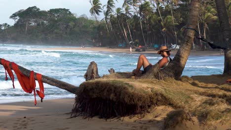 Young-man-with-weaved-hat-laying-against-a-palm-tree,-Handheld-medium-shot