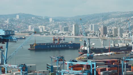 Truck-left-of-cranes,-containers-and-cargo-ships-docked-in-Sea-Port,-Valparaiso-hillside-city-in-background,-Chile
