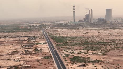 Cinematic-color-graded-aerial-shot-of-silver-car-driving-on-country-road-in-desert-and-big-chimneys-in-distant-background