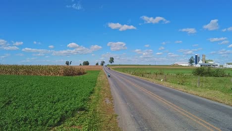 An-Amish-Boy-Riding-a-Scooter,-in-a-Crouched-Position,-Riding-down-a-Country-Road-Hill,-on-a-Sunny-Day