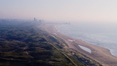 Panning-drone-shot-of-the-lush-dunes-of-Meijendel-and-the-sandy-beach-of-Strand-Wassenaarseslag,-with-the-city-of-Den-Haag-in-the-distance