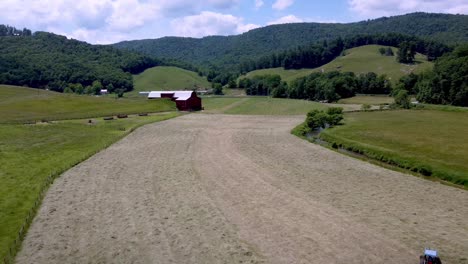 AERIAL-HIGH-SHOT-OF-HAY-BEING-RAKED-WITH-RED-BARN-AND-VALLEY-SETTING-IN-SUMMERTIME-IN-SUGAR-GROVE-NC,-NORTH-CAROLINA-NEAR-BOONE-AND-BLOWING-ROCK-NC,-NORTH-CAROLINA